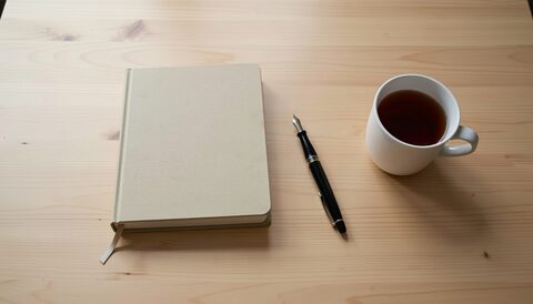 A plain wooden desk seen from above with a closed notebook, a fountain pen and a mug of tea, the surface otherwise empty in even overhead light, no paper in the frame.